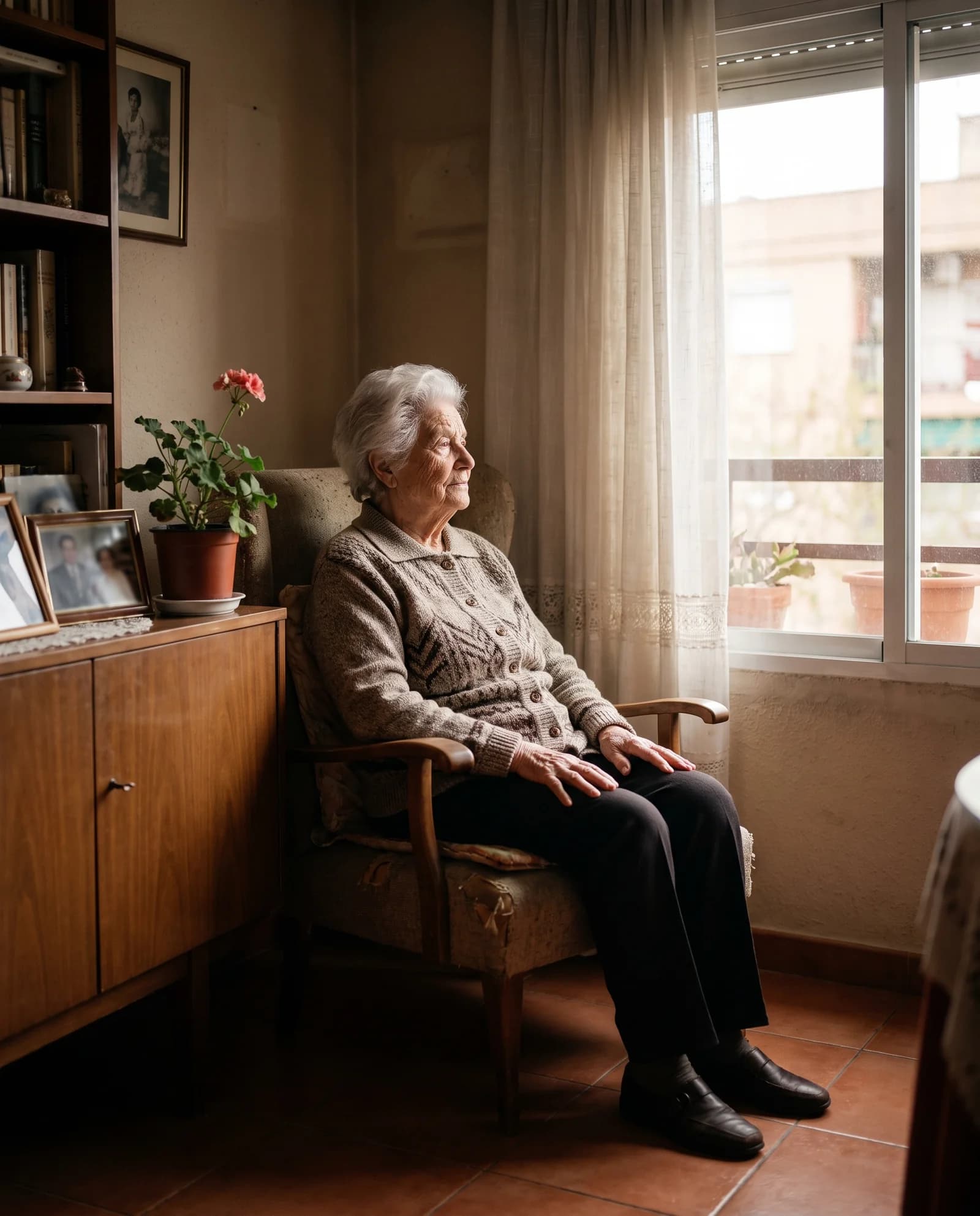 Una mujer mayor sentada junto a la ventana de un salón, con plantas en el balcón.
